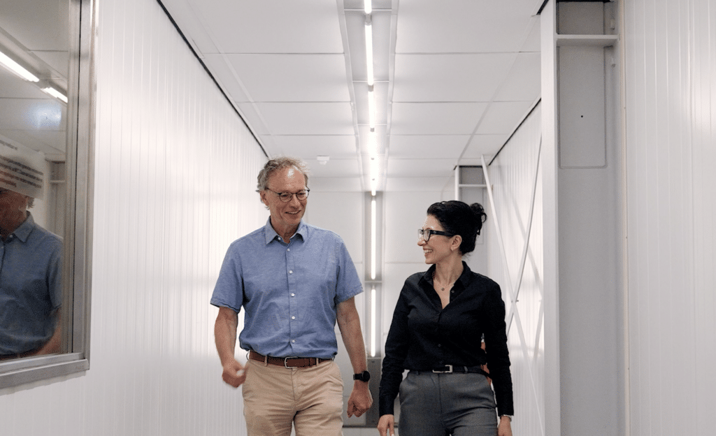 An older man and a woman are walking down a well-lit hallway with white walls. They are smiling and engaged in conversation. The man is wearing a blue short-sleeved shirt and beige trousers, while the woman is dressed in a black blouse and gray pants.