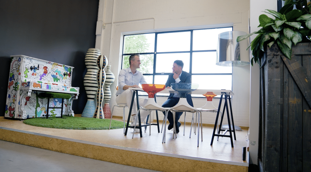 Two men are sitting at a modern table in a creative workspace with large windows. They are engaged in a conversation, smiling at each other. The room features unique decor, including a colorful painted piano, abstract sculptures, and vibrant furniture.